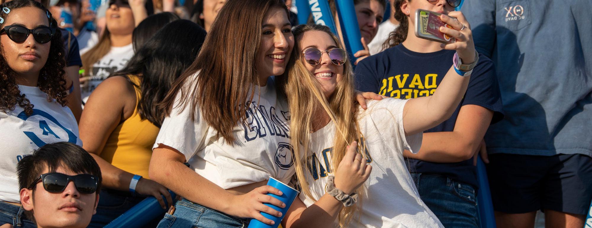Two females students take a selfy in the crowd at a raucous football game
