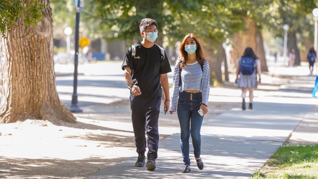 Students with masks walk through the sunlit UC Davis campus.