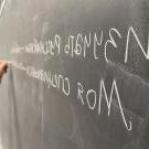A hand writes on a blackboard in white chalk. The words are Russian, printed in Cyrillic.