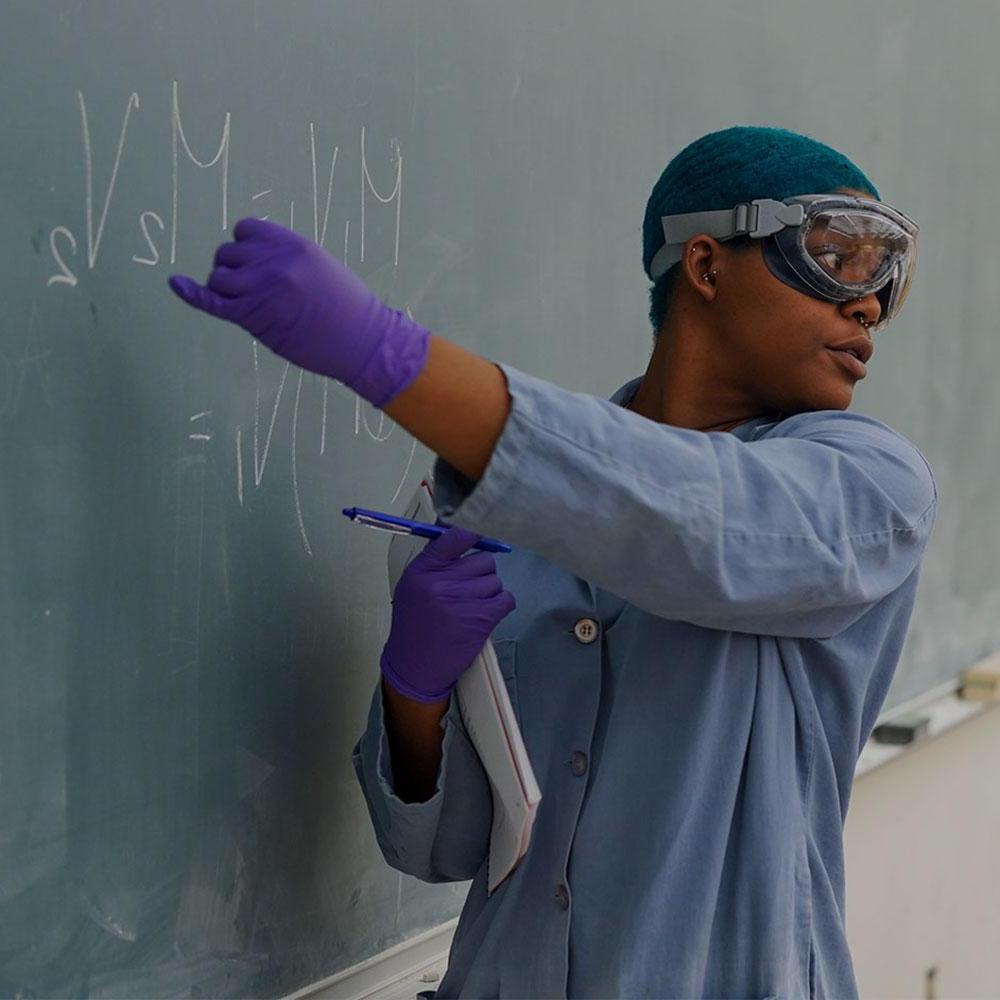 A UC Davis graduate student teaching assistant with short blue hair and wearing a lab coat points to a chemical equation on a blackboard.